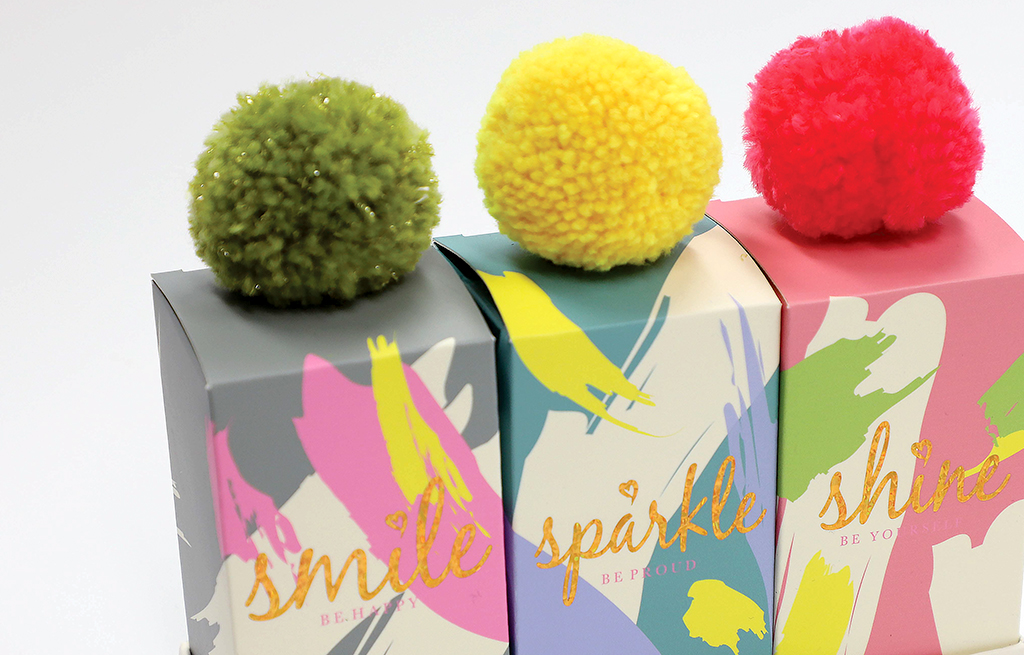 A row of colorful boxes with pom poms on top, in descending order of size. The boxes are blue, green, yellow, and pink, and the pom poms match the colors of the boxes.